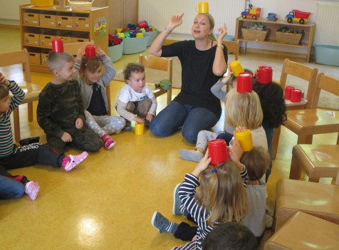 Eine Pädagogin und mehrere Kinder balancieren Becher auf ihrem Kopf