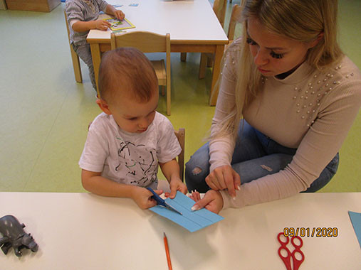Ein Kind schneidet konzentriert, mit Hilfe der Pädagogin, Papierstreifen aus.